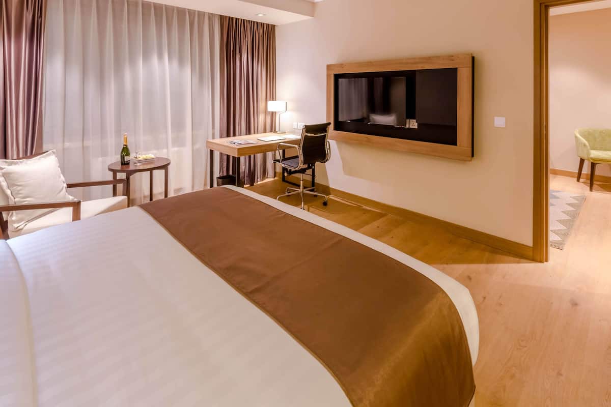 A hotel room with a large bed, a desk with a chair, a television mounted on the wall, and a small table with a bottle and glasses.