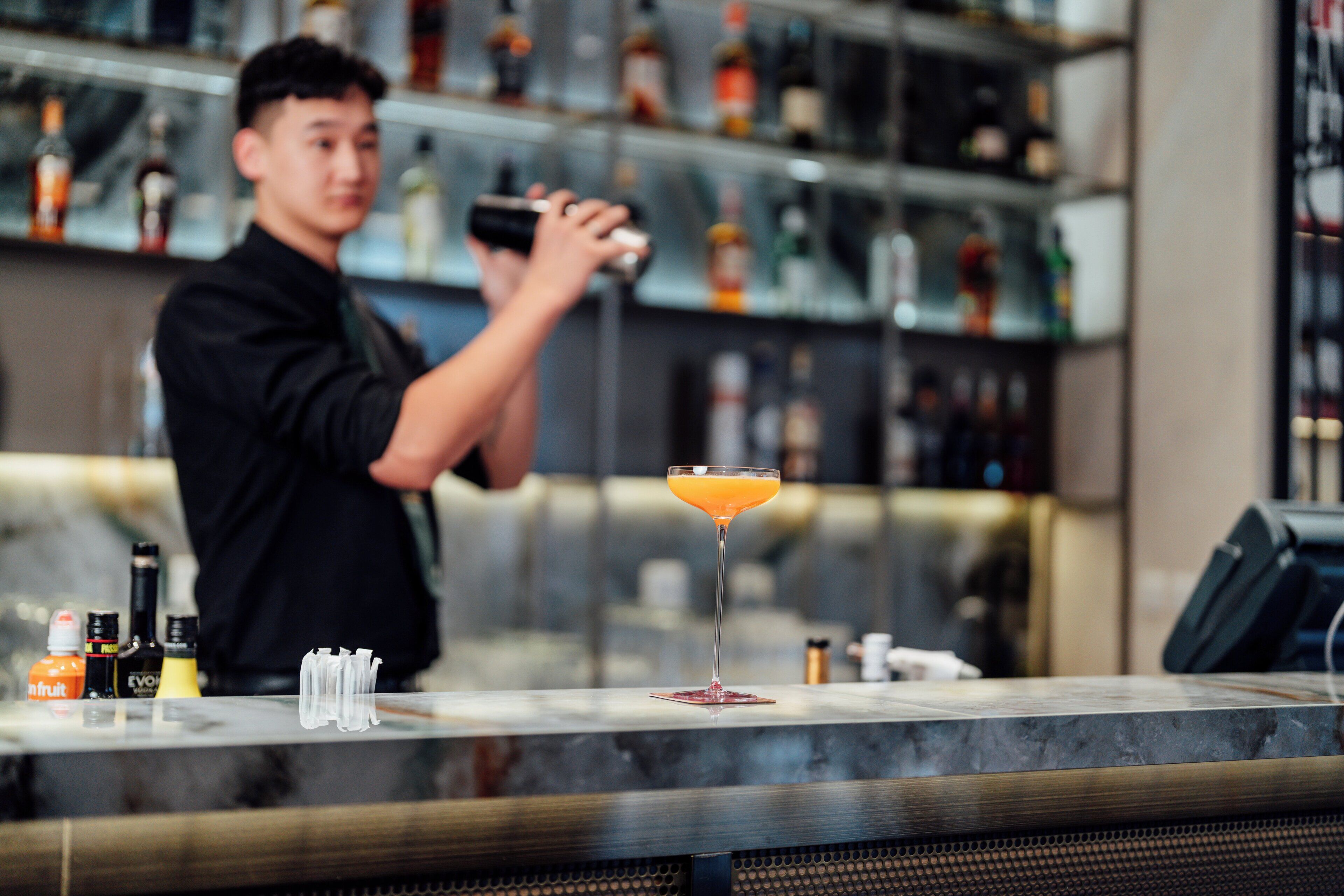 Un barman prépare un cocktail dans un bar où l’on peut voir une variété de bouteilles et un verre à martini posé sur le comptoir.