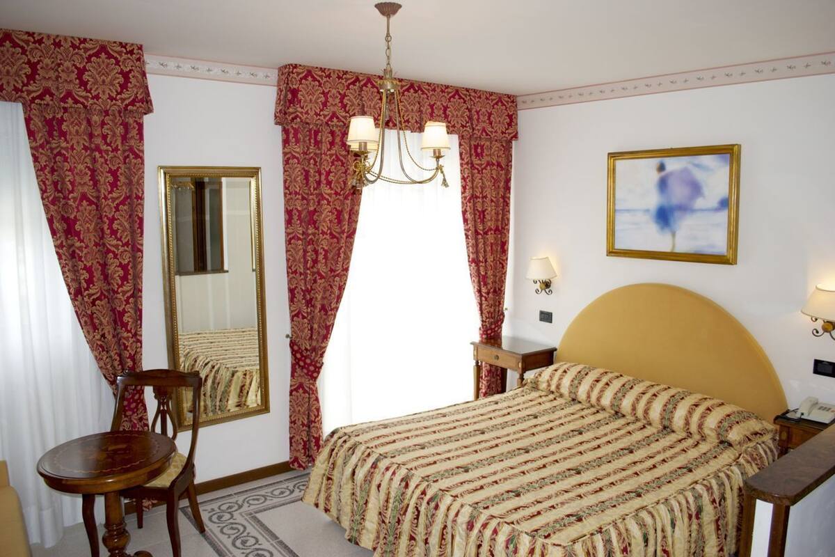 Une chambre d’hôtel avec un lit, une chaise, un bureau, un miroir, un lustre et un tableau au mur.