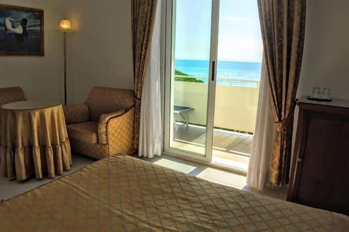 Une chambre d’hôtel avec un lit, une chaise, une petite table et une vue sur la plage, accessible par une porte coulissante.