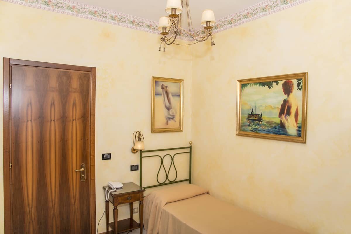 Une pièce avec une porte en bois, un lit avec un cadre en métal vert, un bureau avec une lampe et deux tableaux accrochés au mur.