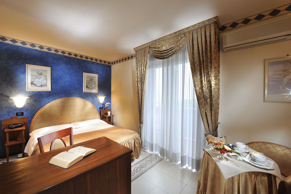 Une chambre d’hôtel avec un grand lit, un bureau en bois, une table à manger dressée pour le petit-déjeuner et deux tableaux encadrés accrochés aux murs.