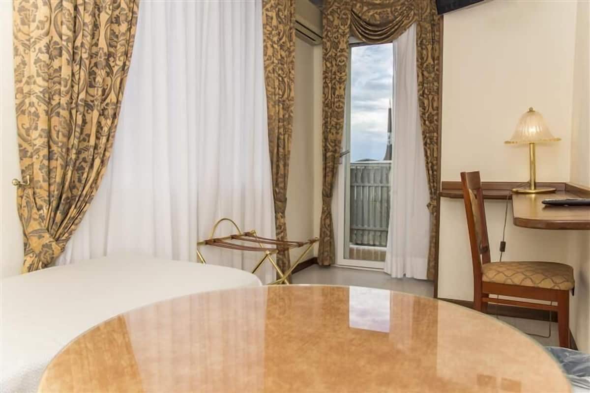 Une chambre d’hôtel avec un lit, une table, une chaise, une lampe et un balcon avec des rideaux.