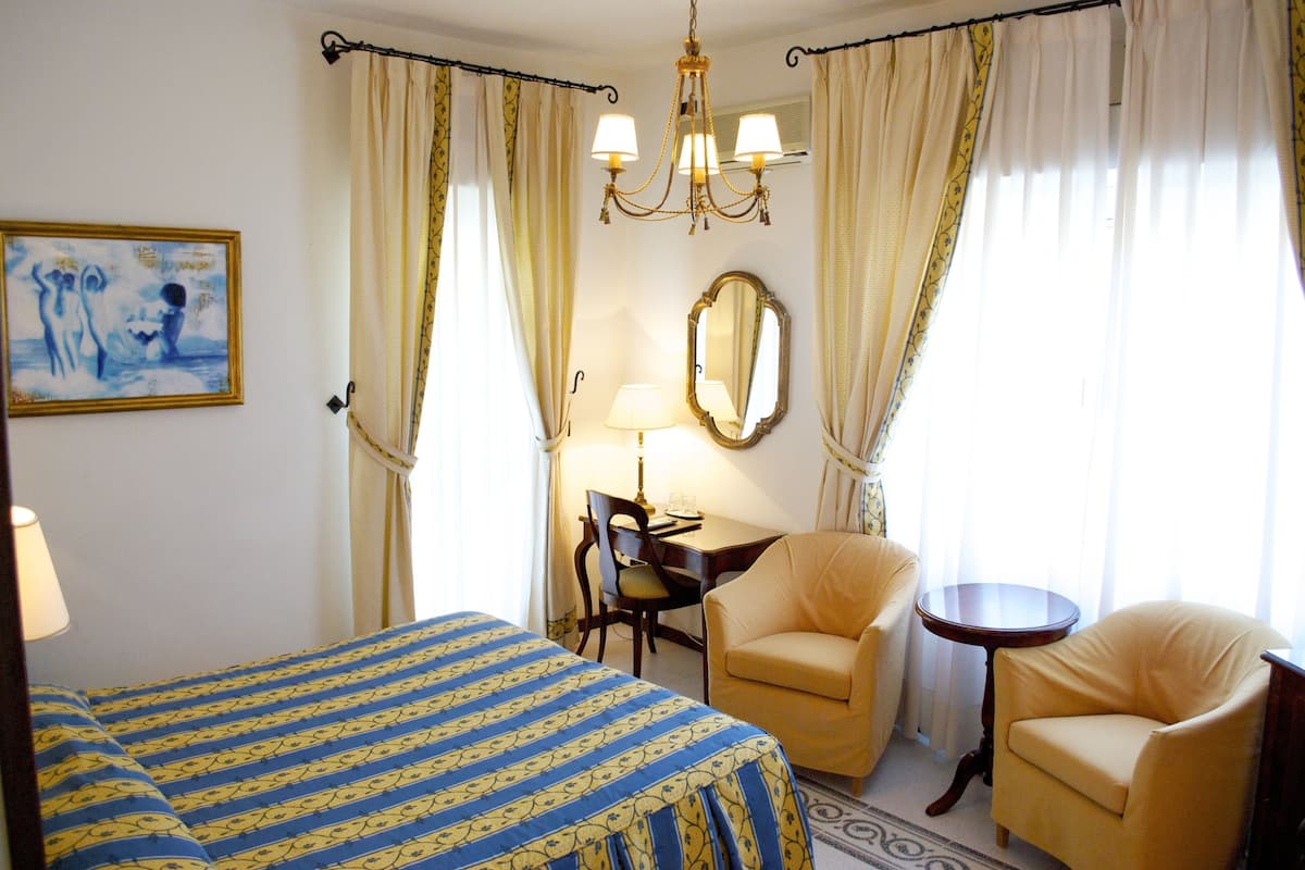 Une chambre d’hôtel comprenant un lit, deux chaises, un bureau avec une lampe, un miroir et un lustre.