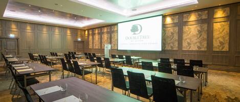 Une salle de conférence équipée de plusieurs tables et chaises, d’un écran de projection affichant le logo « DOUBLETREE by Hilton », et d’un mur en panneaux de bois orné d’un motif floral.