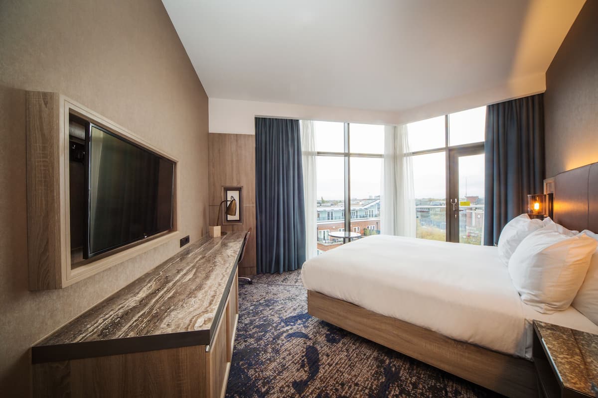 Una habitación de hotel con una cama grande, un escritorio con televisor de pantalla plana, vistas de la ciudad a través de amplias ventanas y un paisaje urbano al exterior.