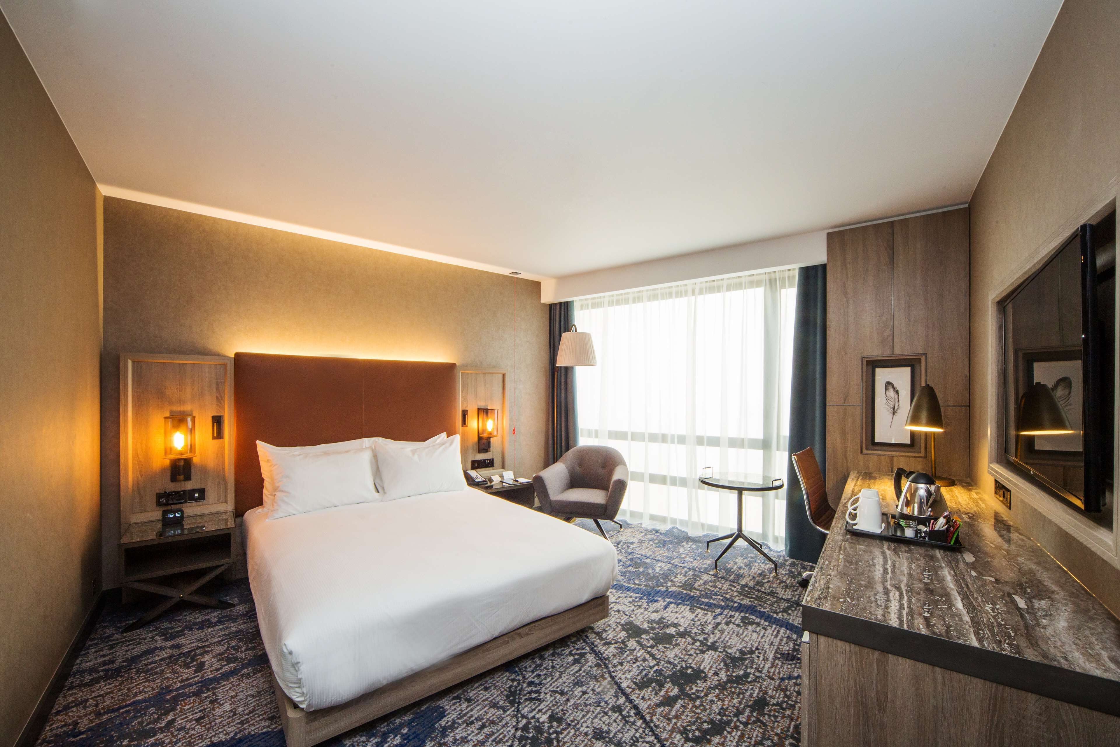 Quarto de hotel com uma cama grande, uma escrivaninha com cadeira, um abajur e uma janela com cortinas.