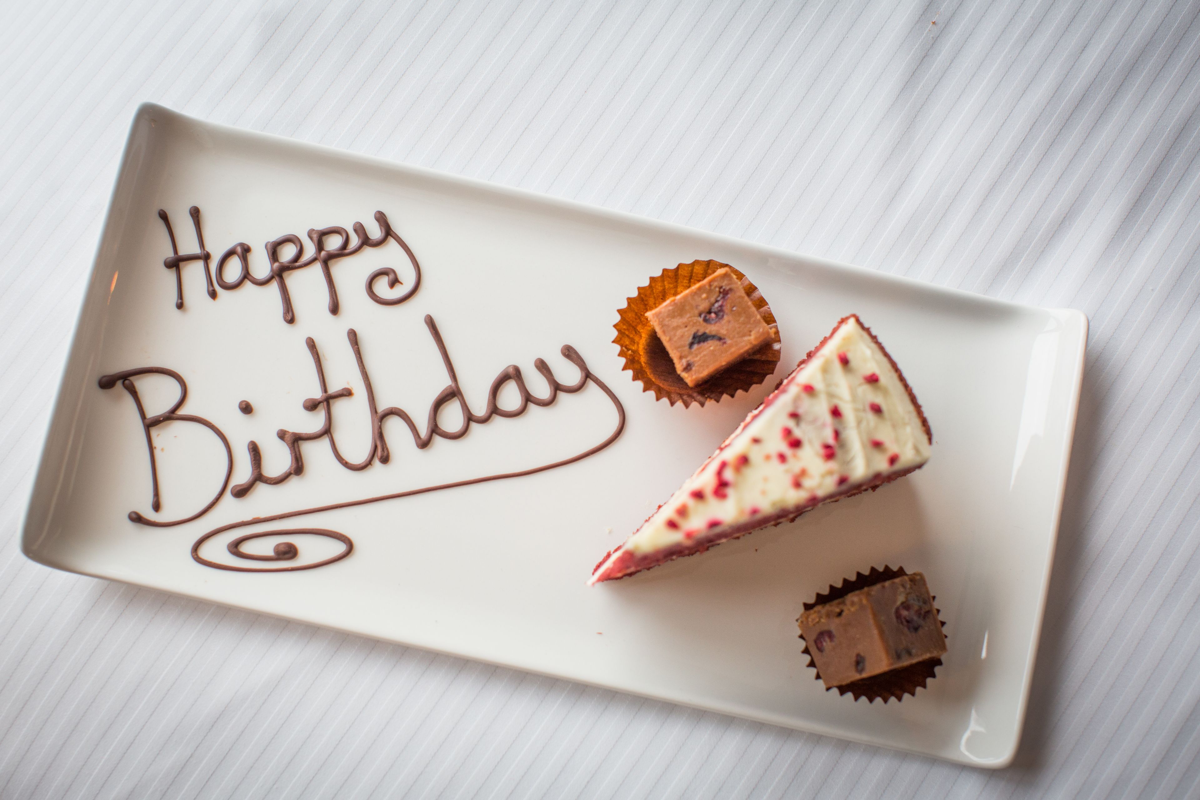 Un plato rectangular blanco con la frase 'Happy Birthday' escrita en glaseado de chocolate, acompañado de tres porciones de pastel y dos bombones cubiertos de chocolate.