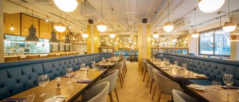 A restaurant with long wooden tables, blue cushioned booths, and pendant lighting.