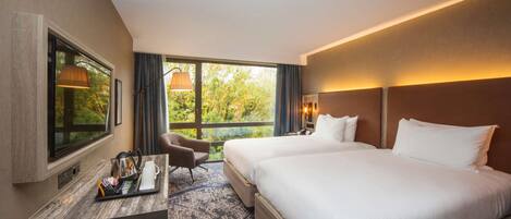 Habitación de hotel con una cama grande, un escritorio con silla, televisor y ventana con vista a árboles.