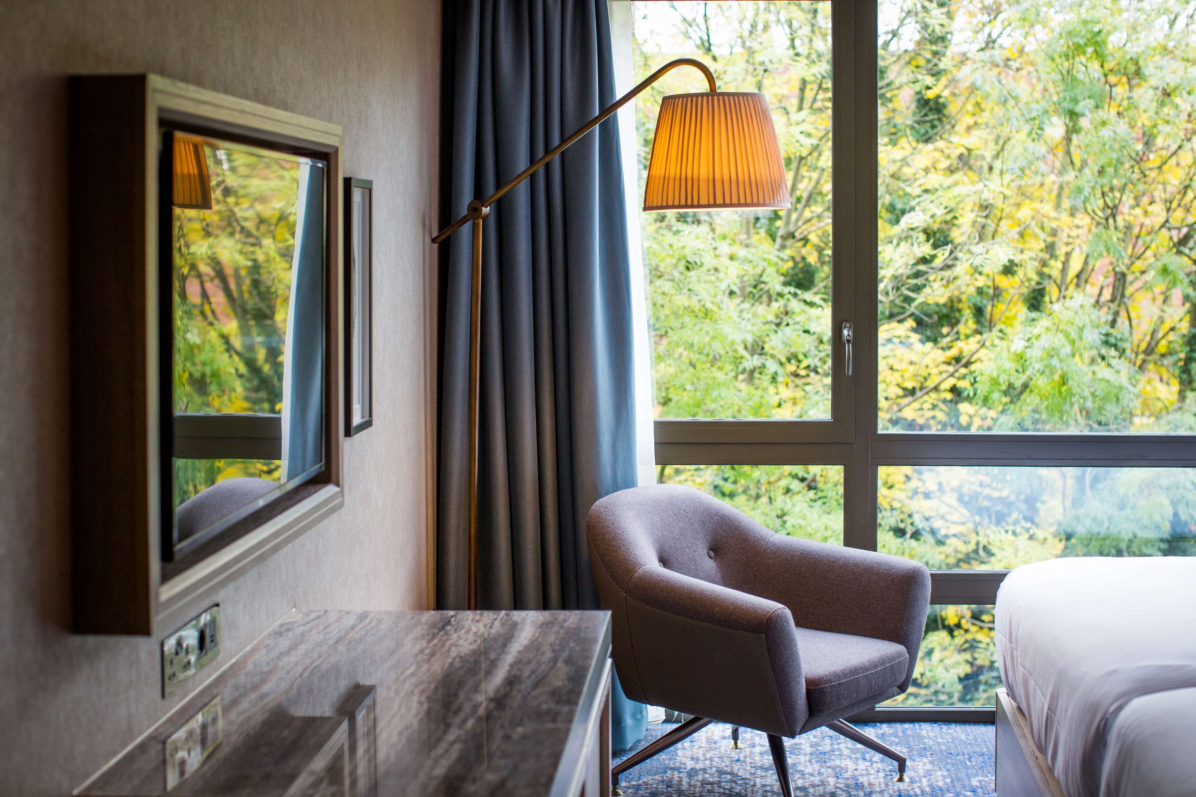 Une chambre d’hôtel avec une grande fenêtre, un lit, une chaise, un bureau et une lampe.