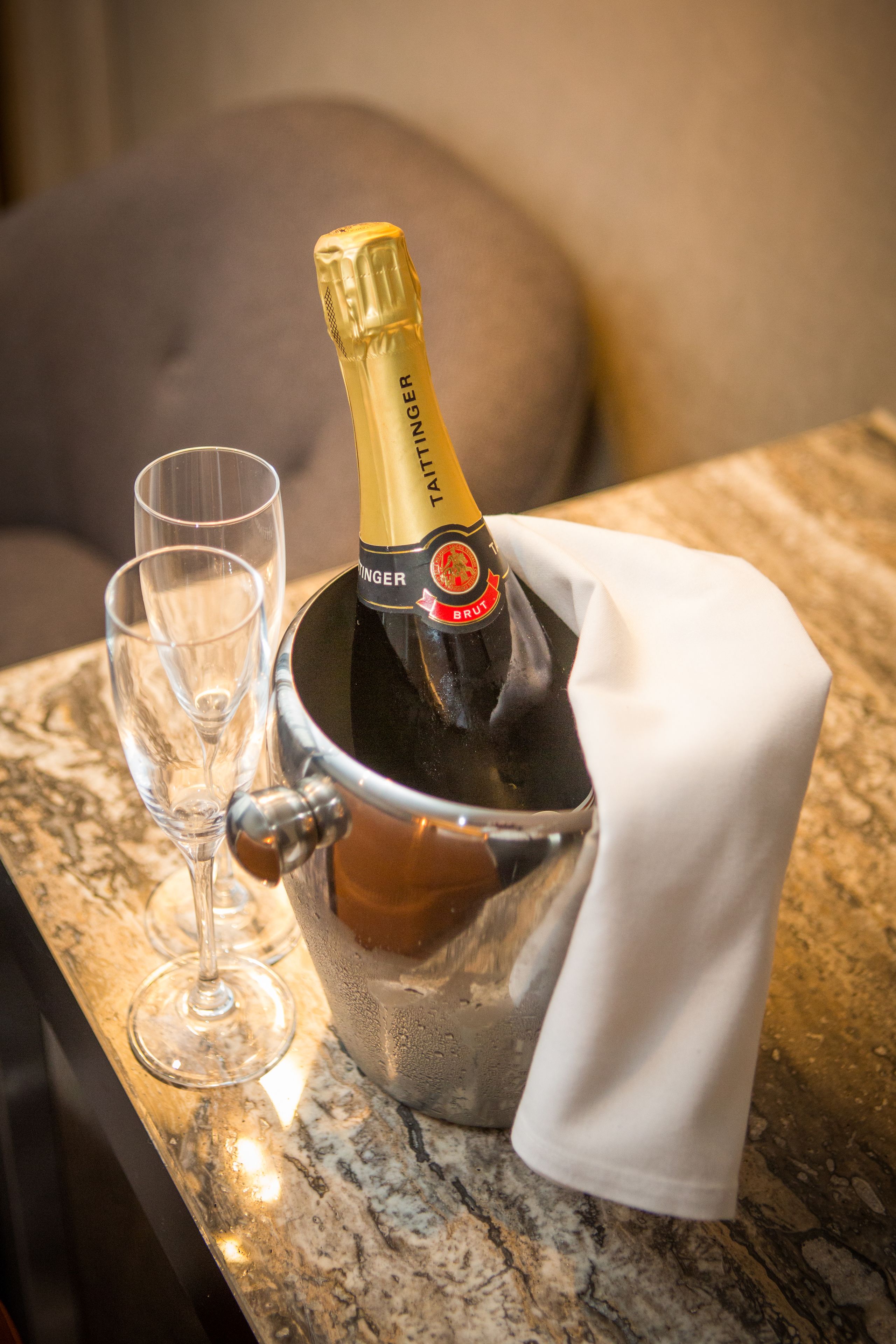 A bottle of champagne in a metal bucket with a white cloth, accompanied by two empty glasses on a marble countertop.