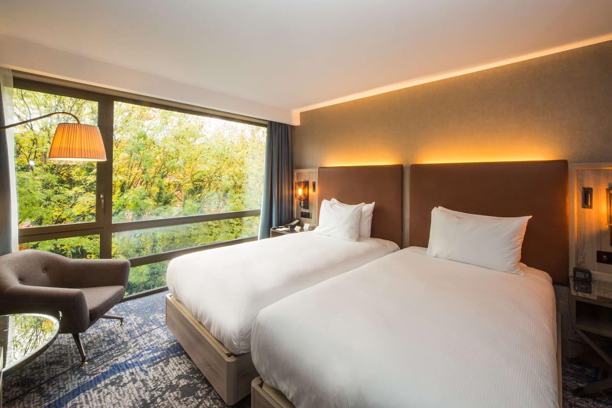 Habitación de hotel con dos camas, una silla, una lámpara y vistas a árboles a través de amplios ventanales.