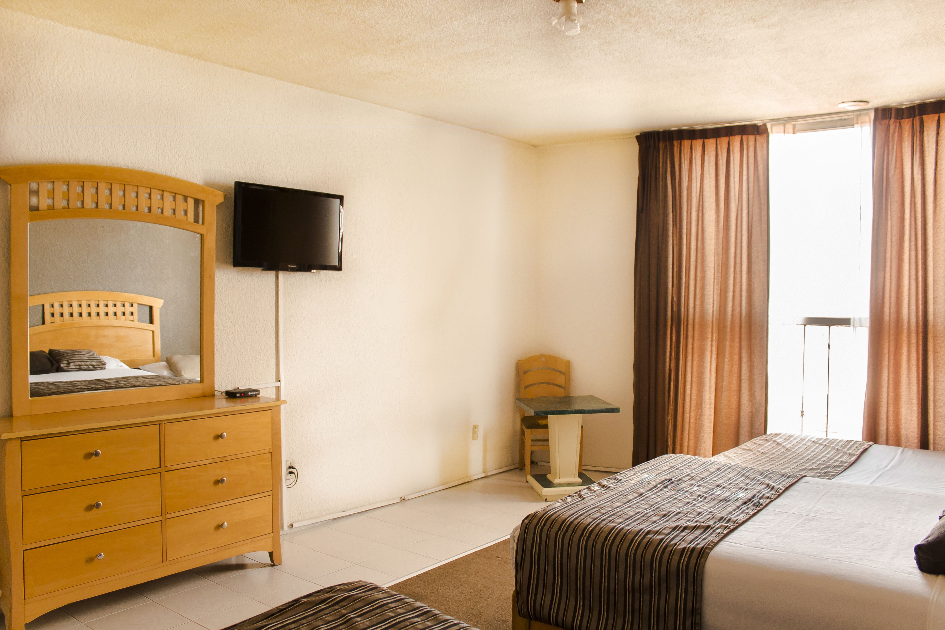 Una habitación de hotel con cama, cómoda, televisor y ventana con cortinas.