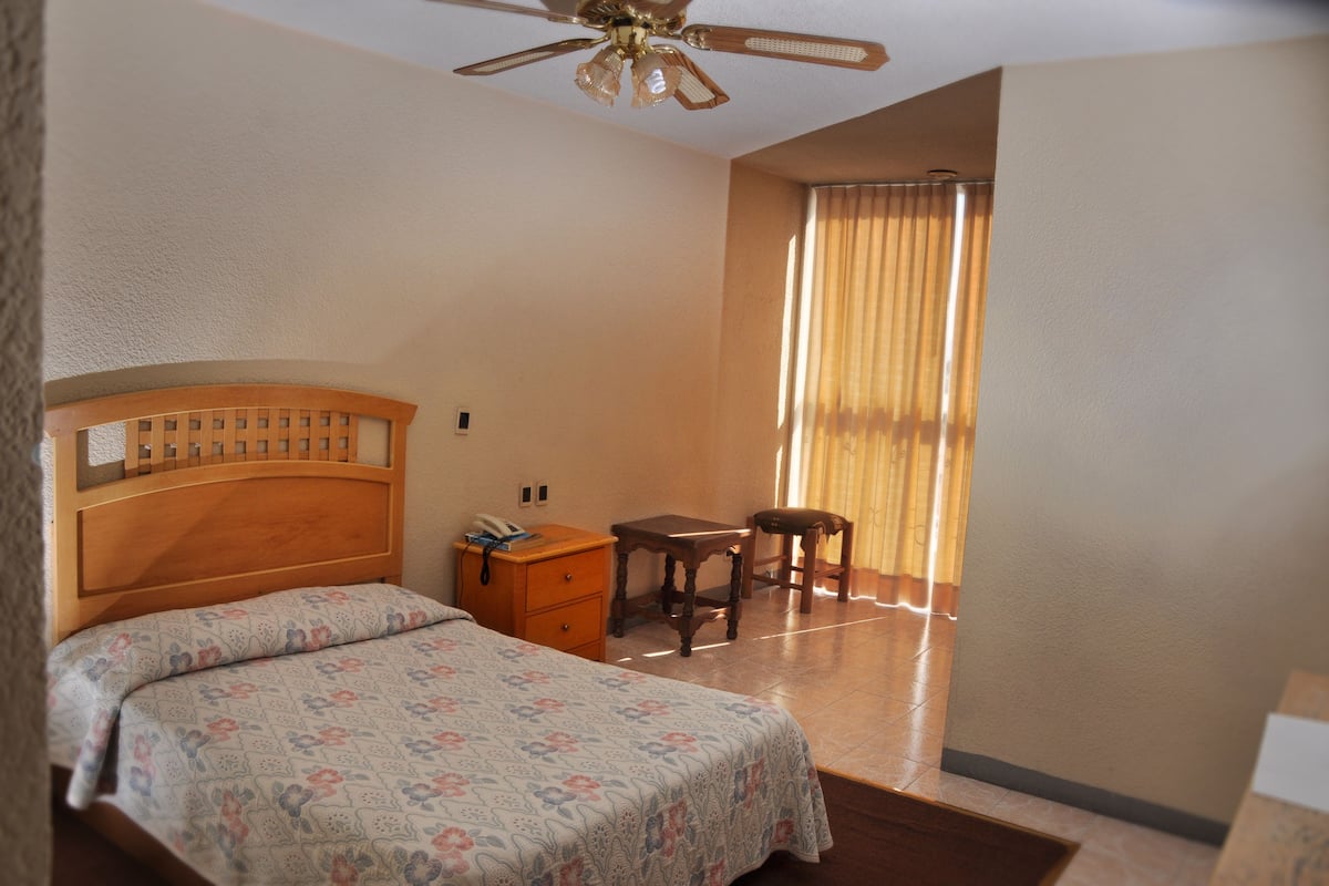Quarto com cama de madeira, mesa de cabeceira, escrivaninha e uma cadeira.