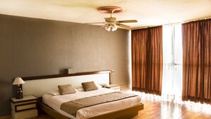 Un dormitorio amplio con una cama grande, una mesita de noche, un ventilador de techo y vistas al exterior a través de cortinas.