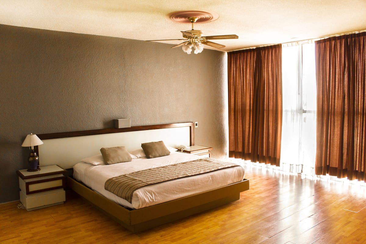 Quarto espaçoso com uma cama grande, mesa de cabeceira, ventilador de teto e vista para o exterior através de cortinas.