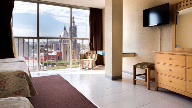 Habitación de hotel con balcón grande, una cama, cómoda de madera, una silla y televisor fijado a la pared.