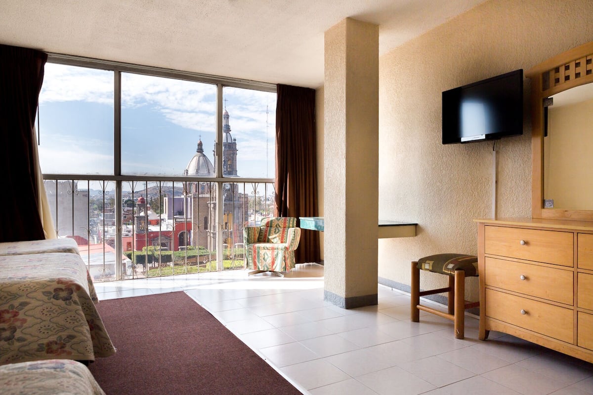 Quarto de hotel com uma varanda grande, uma cama, penteadeira de madeira, uma cadeira e uma TV fixada na parede.