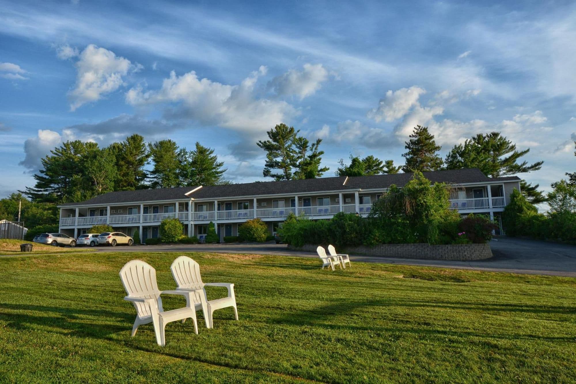 A motel with a two-story structure, a parking area, and a grassy lawn with two Adirondack chairs.