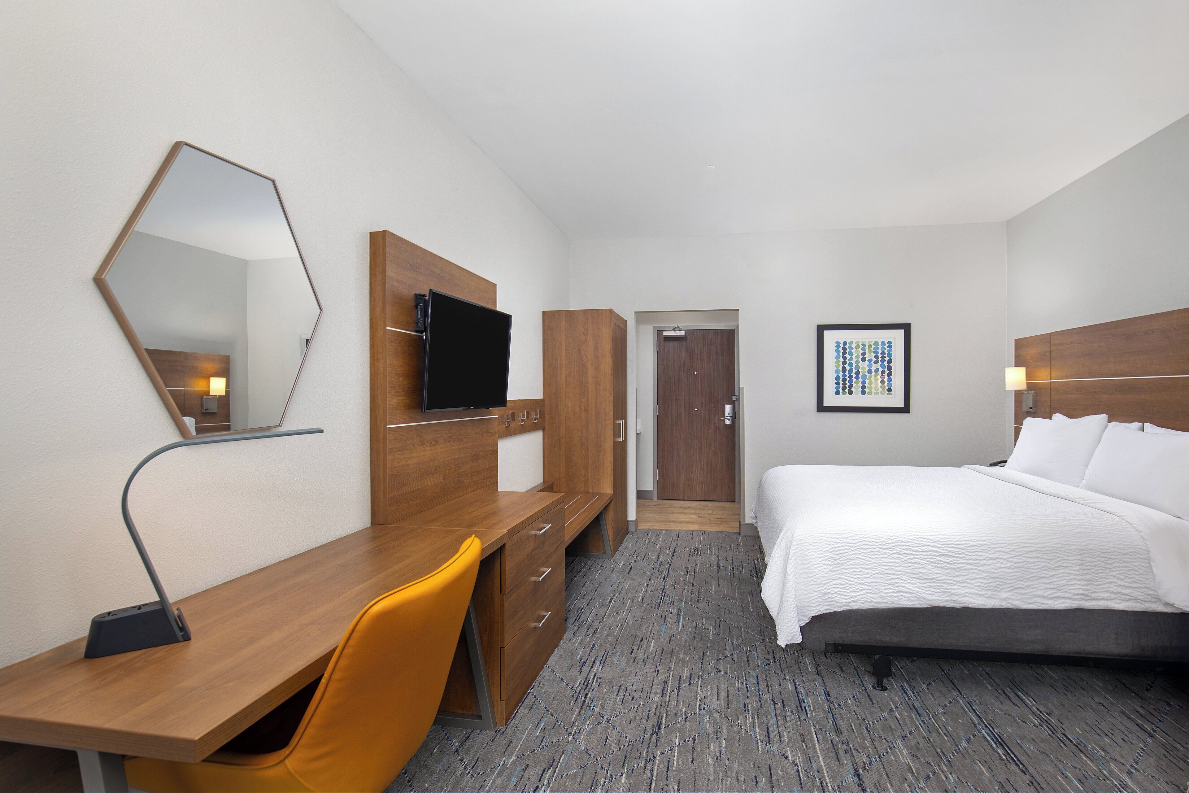 A hotel room with a wooden desk, a chair, a bed, a flat-screen TV, a framed artwork, and a door leading to another room.