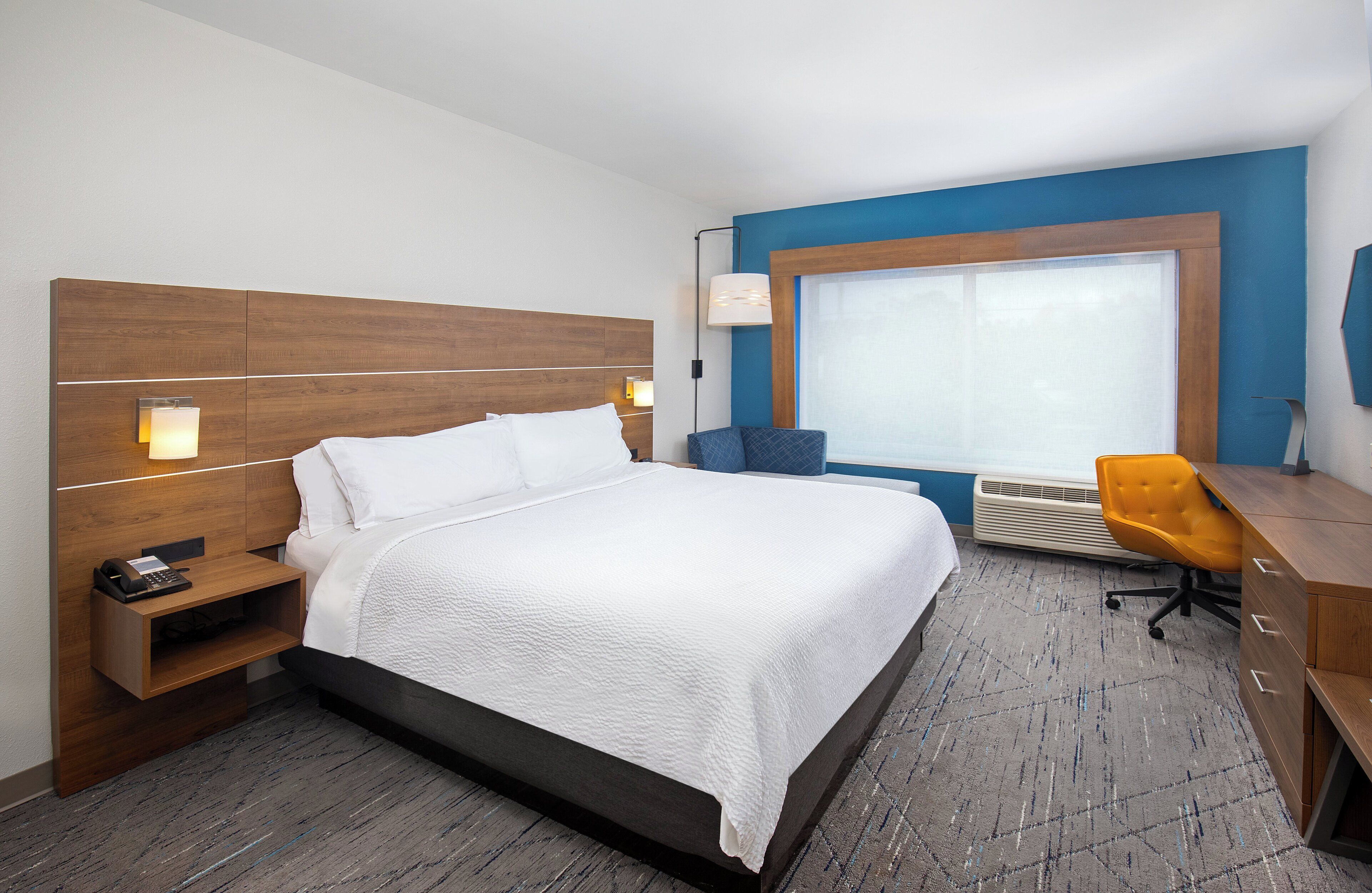 A hotel room with a large bed, a desk with a chair, a window with blinds, and a wall-mounted lamp.