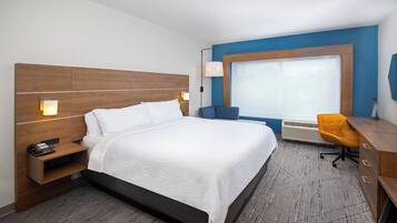 A hotel room with a large bed, a desk with a chair, a window with blinds, and a wall-mounted lamp.