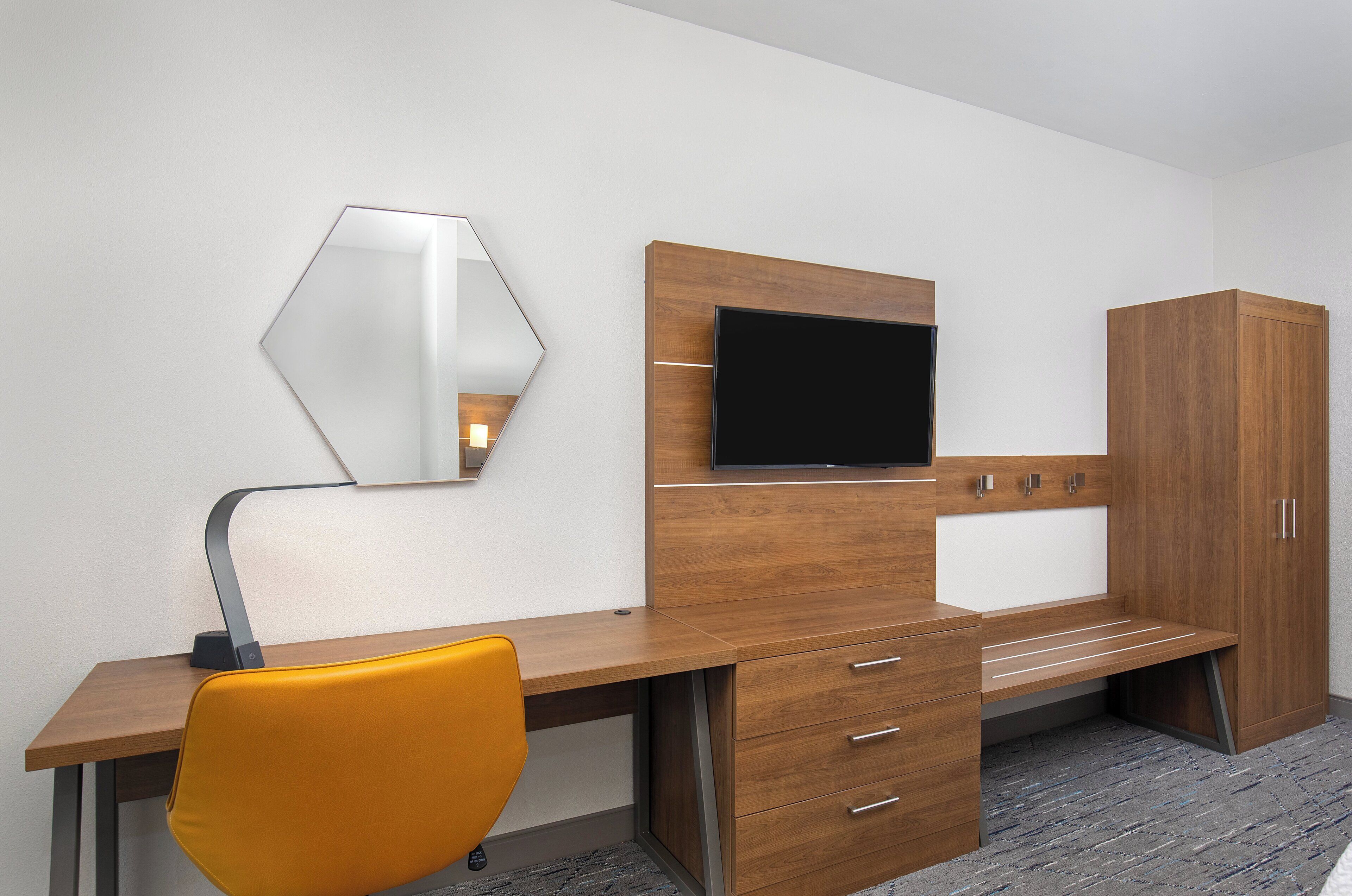 A modern hotel room with a wooden desk, a flat-screen TV, a mirror, and a wardrobe.