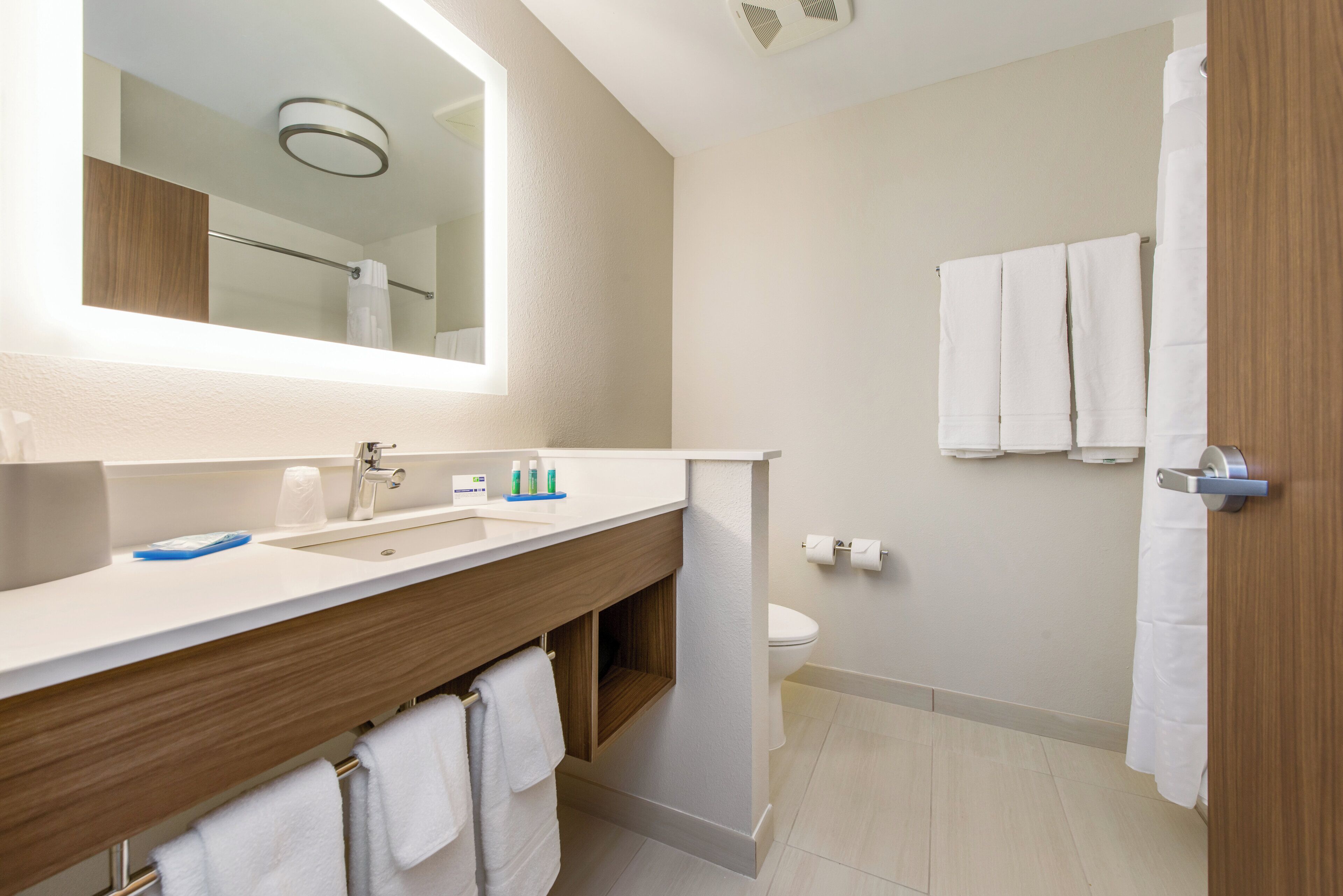 Une salle de bain moderne avec un grand miroir, un lavabo et des toilettes. Des serviettes sont accrochées au mur et il y a un rideau de douche.
