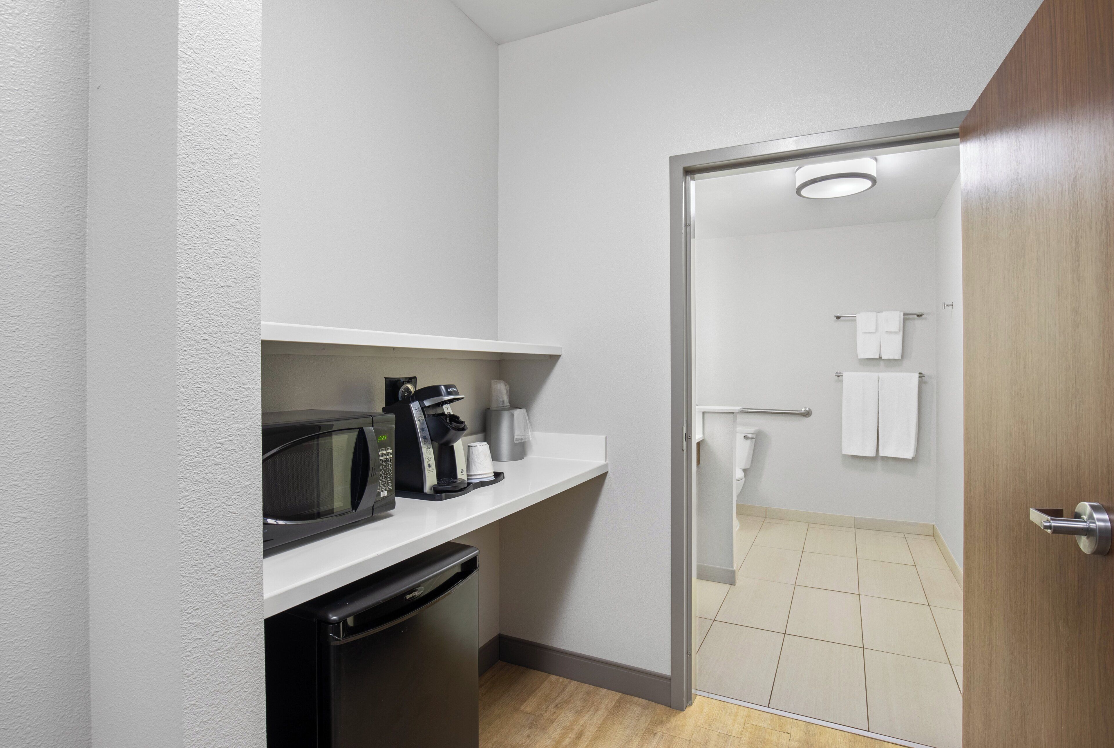 A compact kitchenette with a microwave, coffee maker, and toaster. A door leads to a bathroom with a toilet and towel rack.