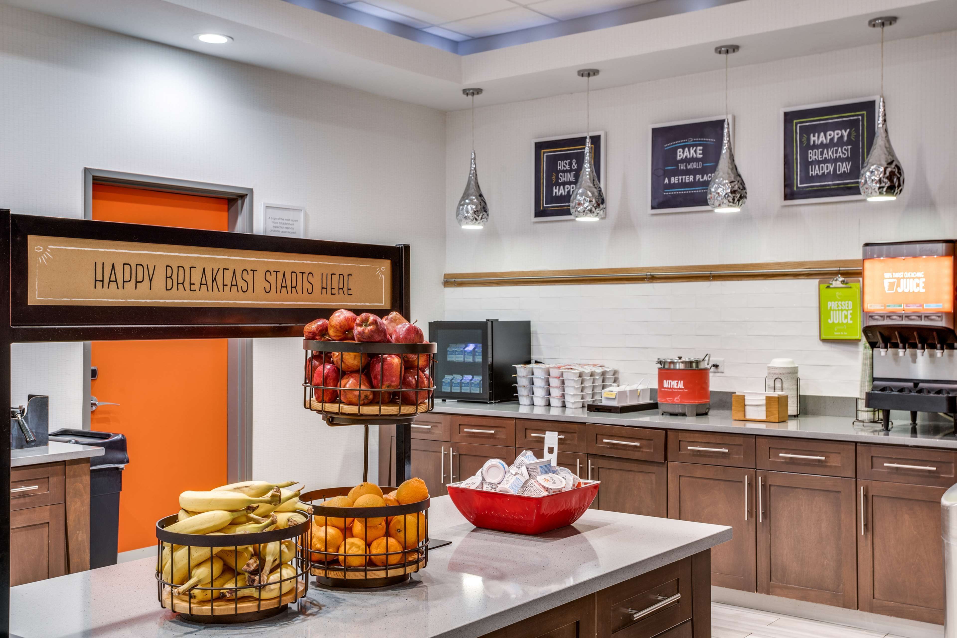 A breakfast area with a sign that reads "HAPPY BREAKFAST STARTS HERE," a fruit basket, a bowl of bread, and a juice dispenser.