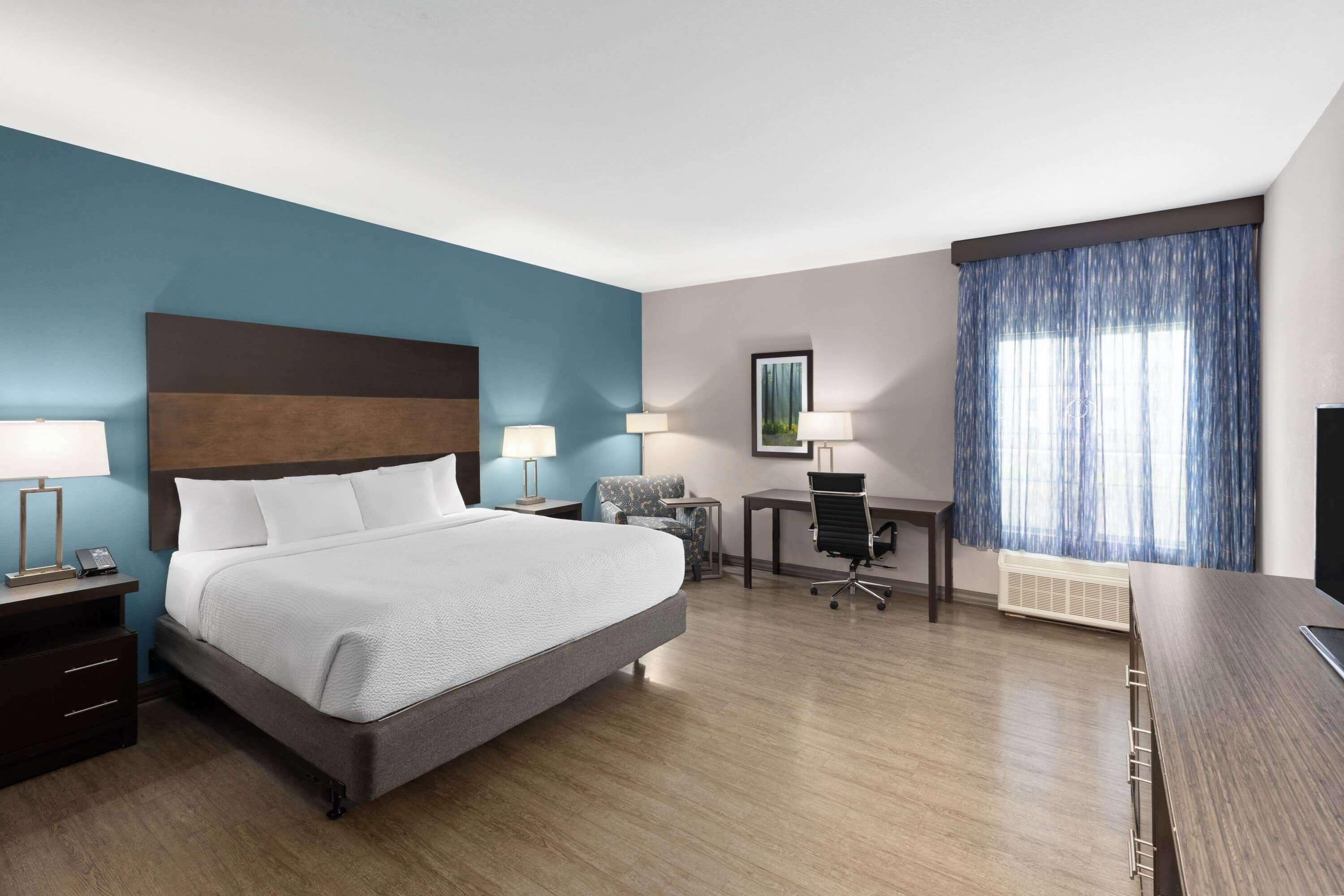 A hotel room with a large bed, two bedside tables with lamps, a desk with a chair, a window with curtains, and a mounted TV.
