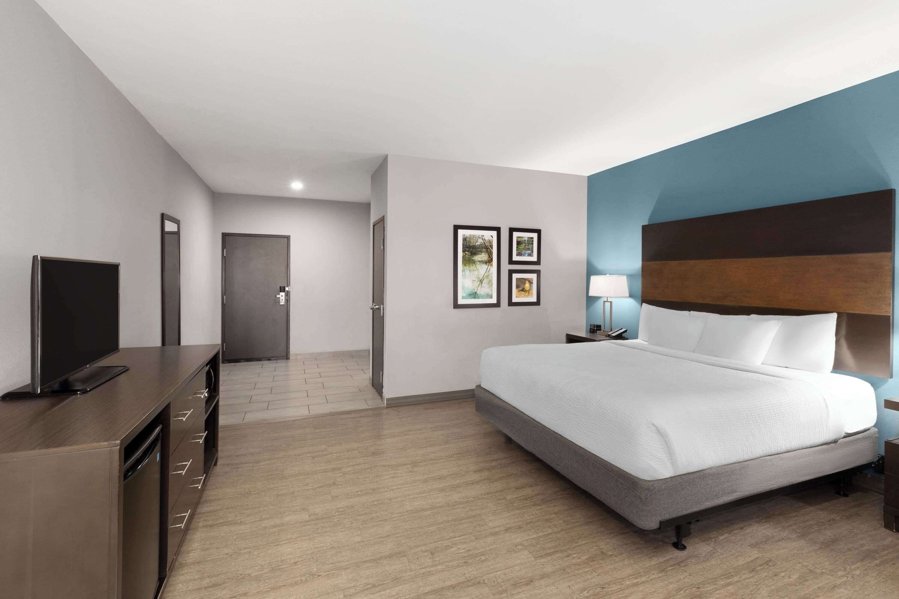 Ein modernes Hotelzimmer mit einem großen Bett, einem Flachbildfernseher, einem Schreibtisch mit Schubladen und einer Tür zu einem weiteren Raum.