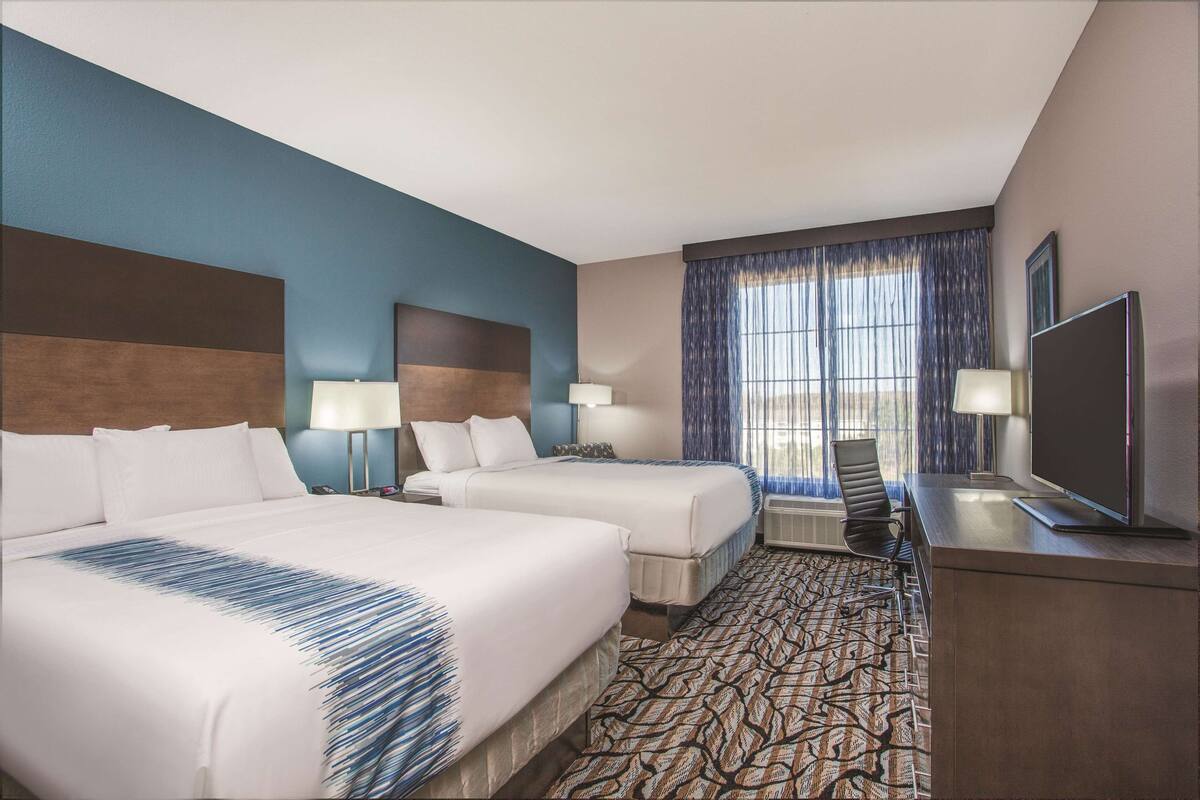 A hotel room with two beds, a desk with a TV, and a large window with curtains.