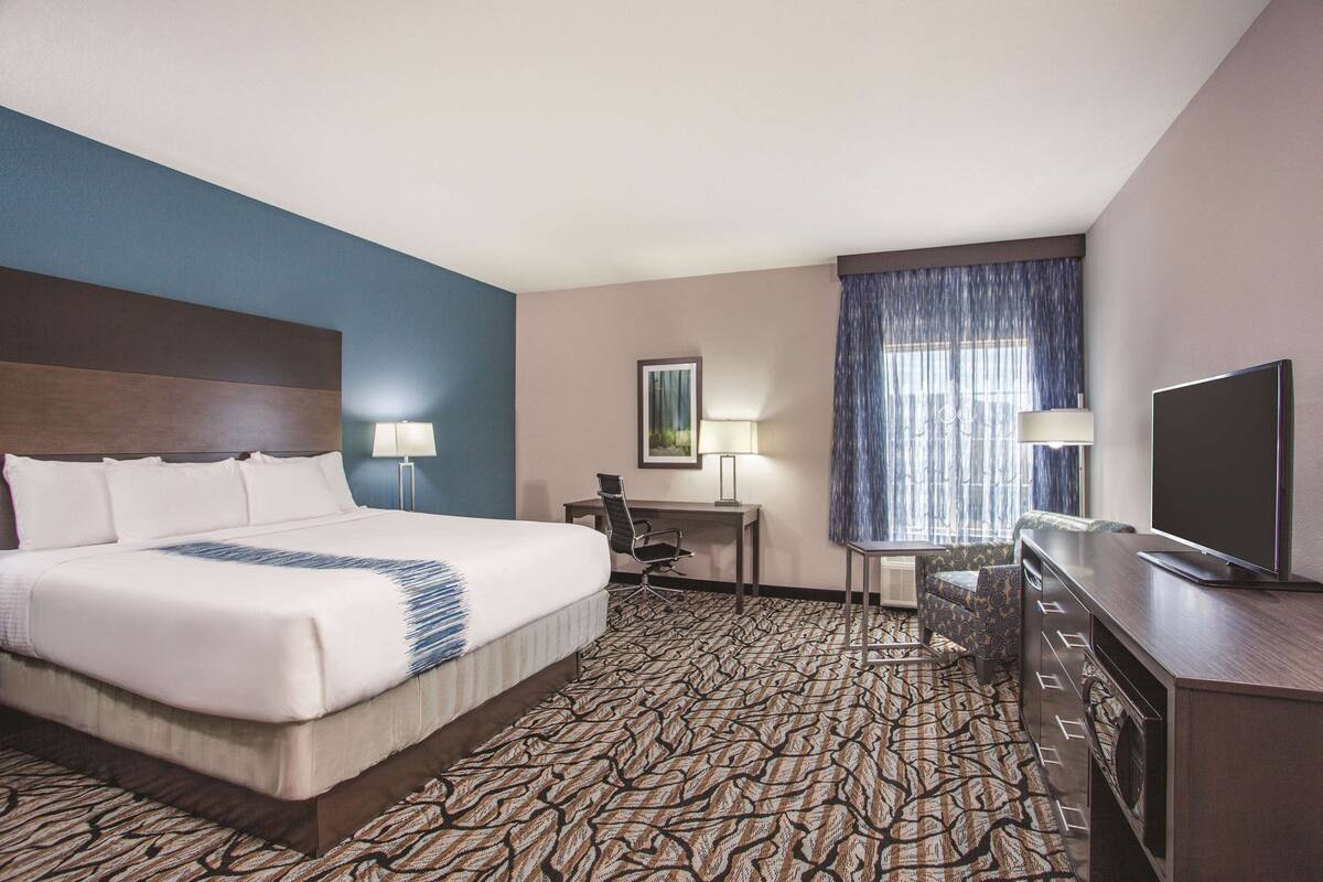 A hotel room with a large bed, a desk, a chair, a television, and a patterned carpet.
