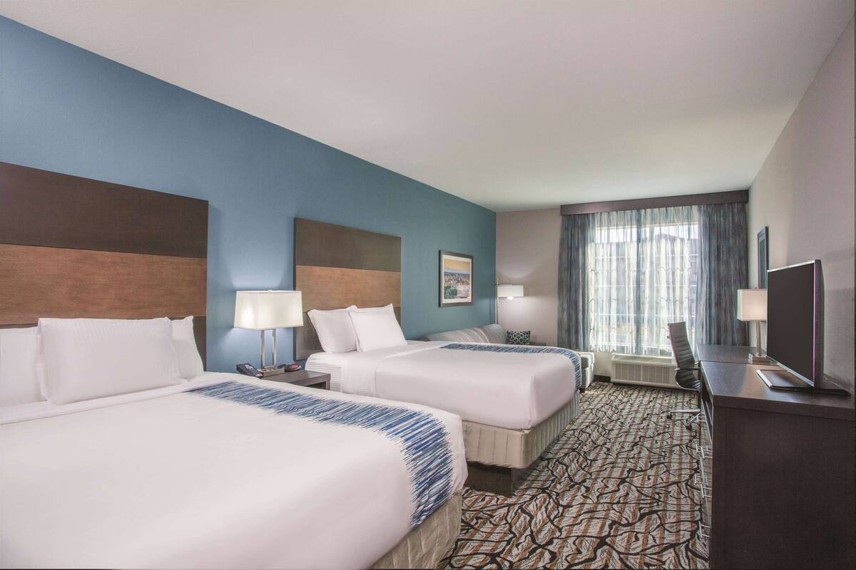 A hotel room with two beds, a desk, a TV, and a large window with curtains.