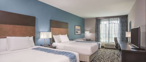 A hotel room with two beds, a desk, a TV, and a large window with curtains.