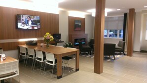 A modern living room with a wooden dining table, white chairs, a flat-screen TV mounted on the wall, a fireplace, and a large window. - Microtel Inn & Suites By Wyndham Binghamton (Binghamton)