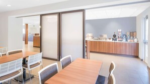 A modern dining area with wooden tables and white chairs, a kitchen counter with various appliances, and a view of the adjacent room. - Microtel Inn & Suites By Wyndham Binghamton (Binghamton)