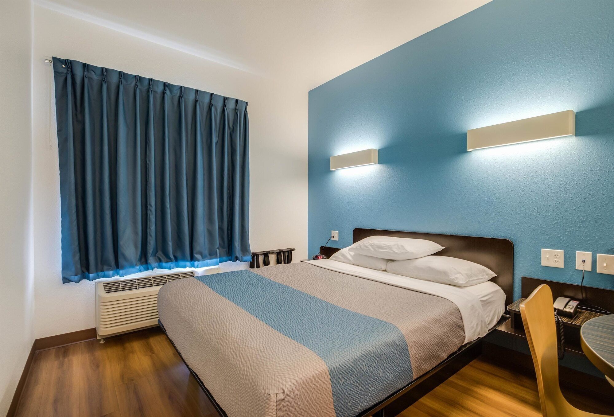 Una habitación de hotel con cama, escritorio y silla, ventana con cortinas e iluminación en la pared.