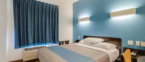 Una habitación de hotel con cama, escritorio y silla, ventana con cortinas e iluminación en la pared.