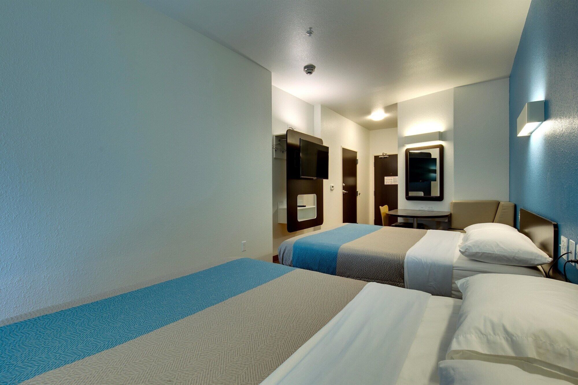 A hotel room with two beds, a desk, a chair, a mirror, and a TV mounted on the wall.