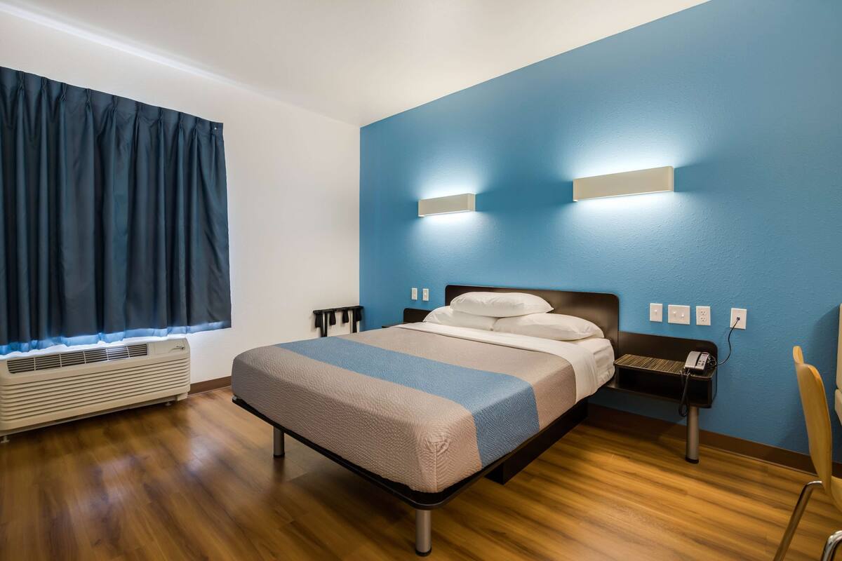 Habitación de hotel con cama, mesitas de noche, lámpara de pared, equipo de aire acondicionado y ventana con cortinas.