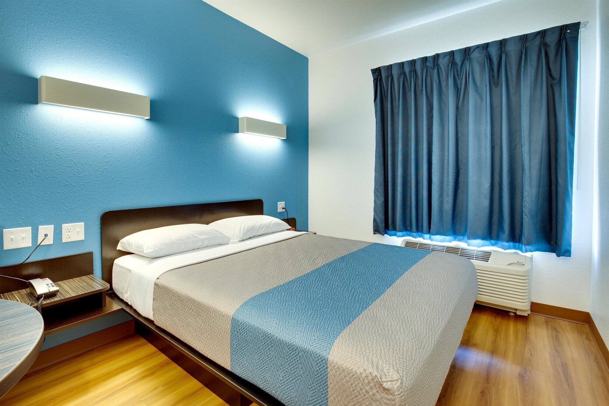 Una habitación de hotel con una cama, mesita de noche y un ventanal con cortinas.