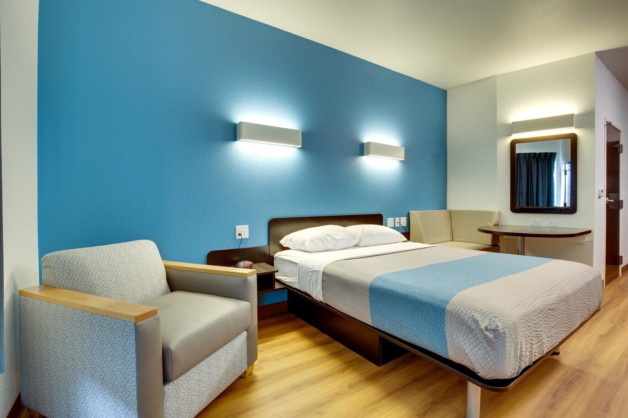 Una habitación de hotel con una cama, una silla, una mesita de noche, un espejo y una lámpara fijada a la pared.