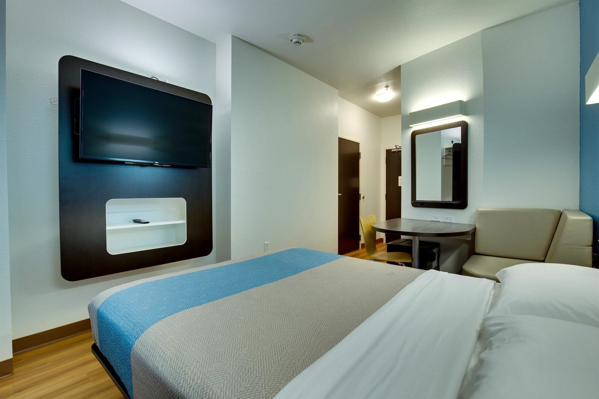 A hotel room with a bed, a desk, a chair, a mirror, and a TV mounted on the wall.