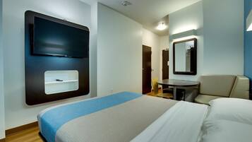A hotel room with a bed, a desk, a chair, a mirror, and a TV mounted on the wall.