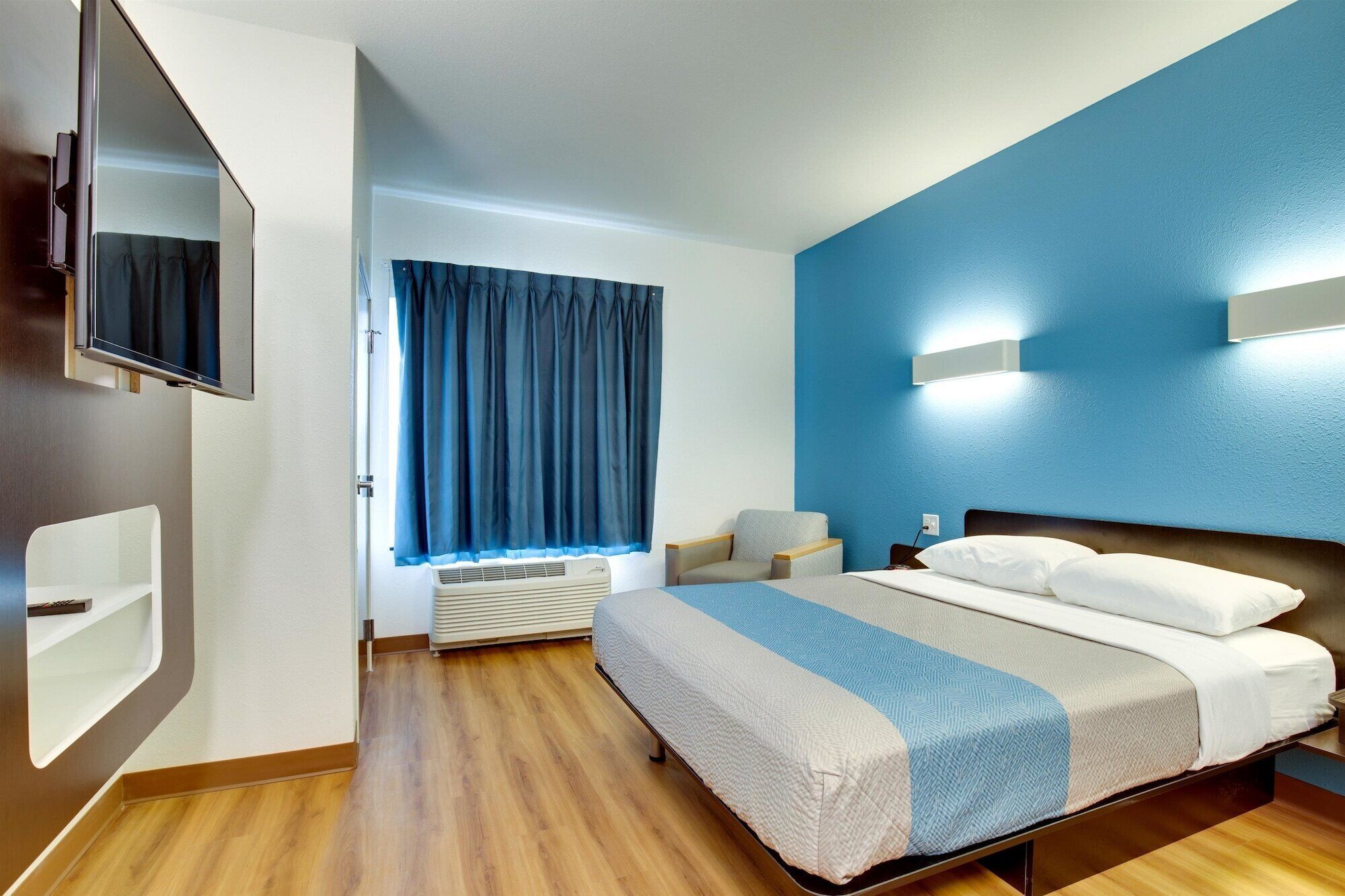 A modern hotel room with a large bed, a flat-screen TV mounted on the wall, a window with blue curtains, and a built-in air conditioning unit.