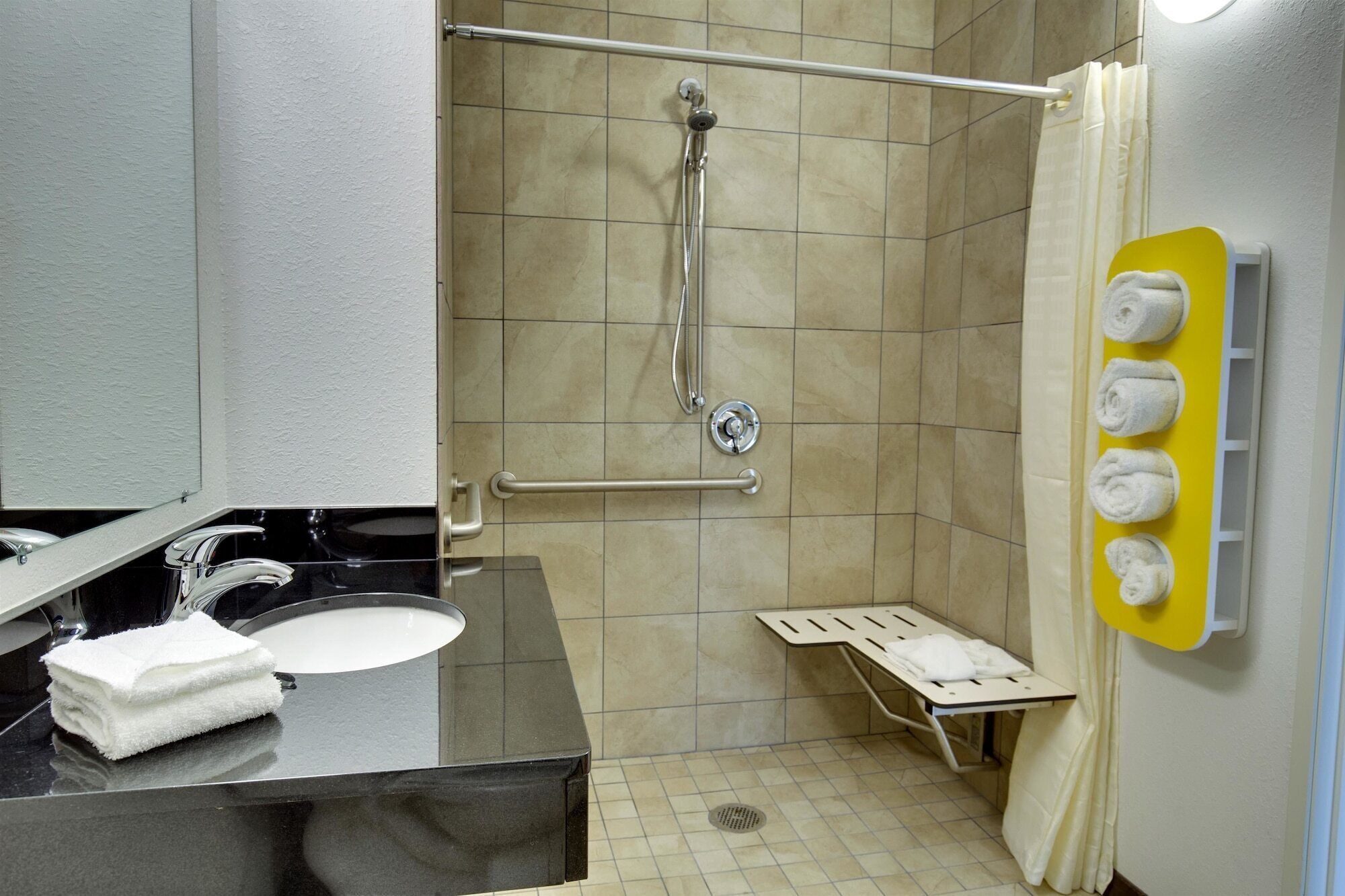 A bathroom with a sink, mirror, towel dispenser, shower, and grab bars.