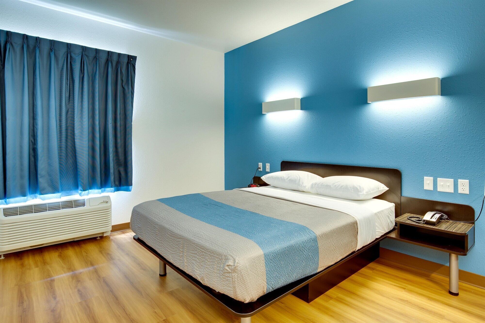 Habitación de hotel con cama, mesita de noche, lámpara, ventana con cortinas y una lámpara de pared.